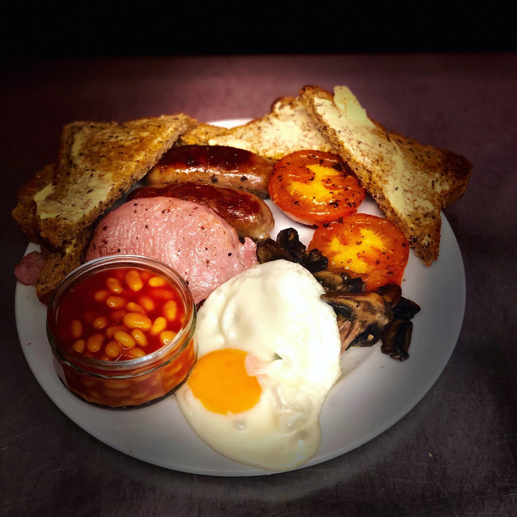 A cooked breakfast with ham, sausages, egg, beans and toast