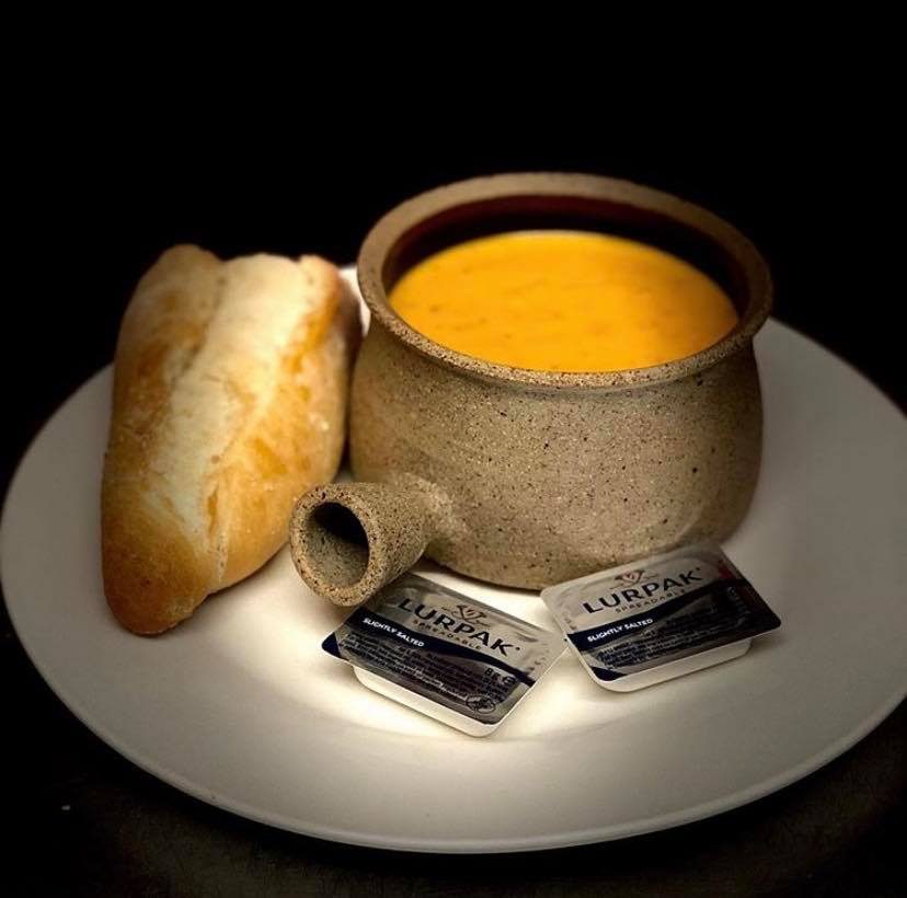 A pot of orange soup with a bread roll and butter